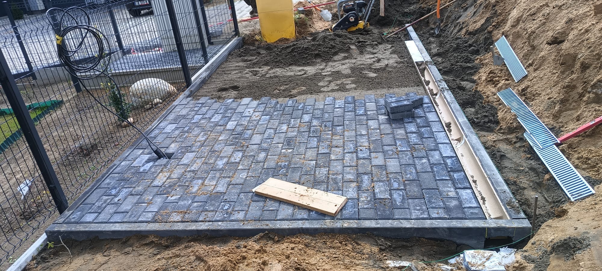 Fragment układanej szarej kostki brukowej na podsypce piaskowej, widoczne korytka odwadniające, ubijarka, ogrodzenie panelowe i narzędzia budowlane.