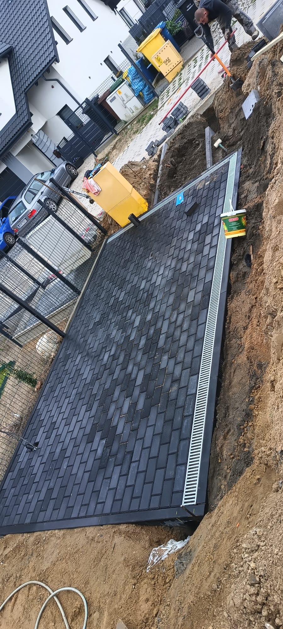 Ciemnoszara kostka brukowa ułożona na podwórku, obok widoczny drenaż liniowy, częściowo wykopana ziemia, w tle dom i pracownik z łopatą.