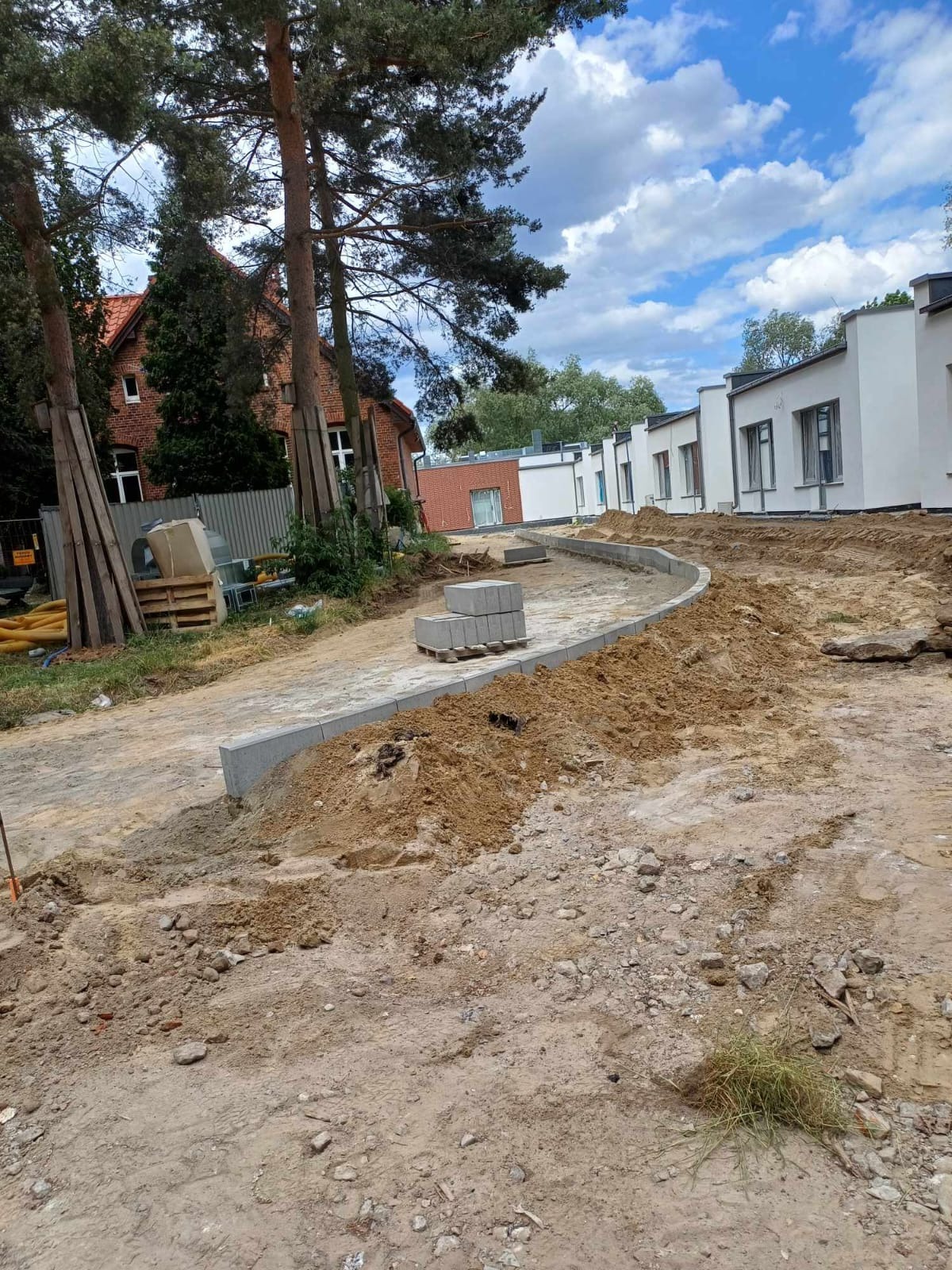 Przygotowanie podłoża pod ścieżkę z obrzeżami, widoczne sterty ziemi, paleta z kostką betonową i budynki w tle.