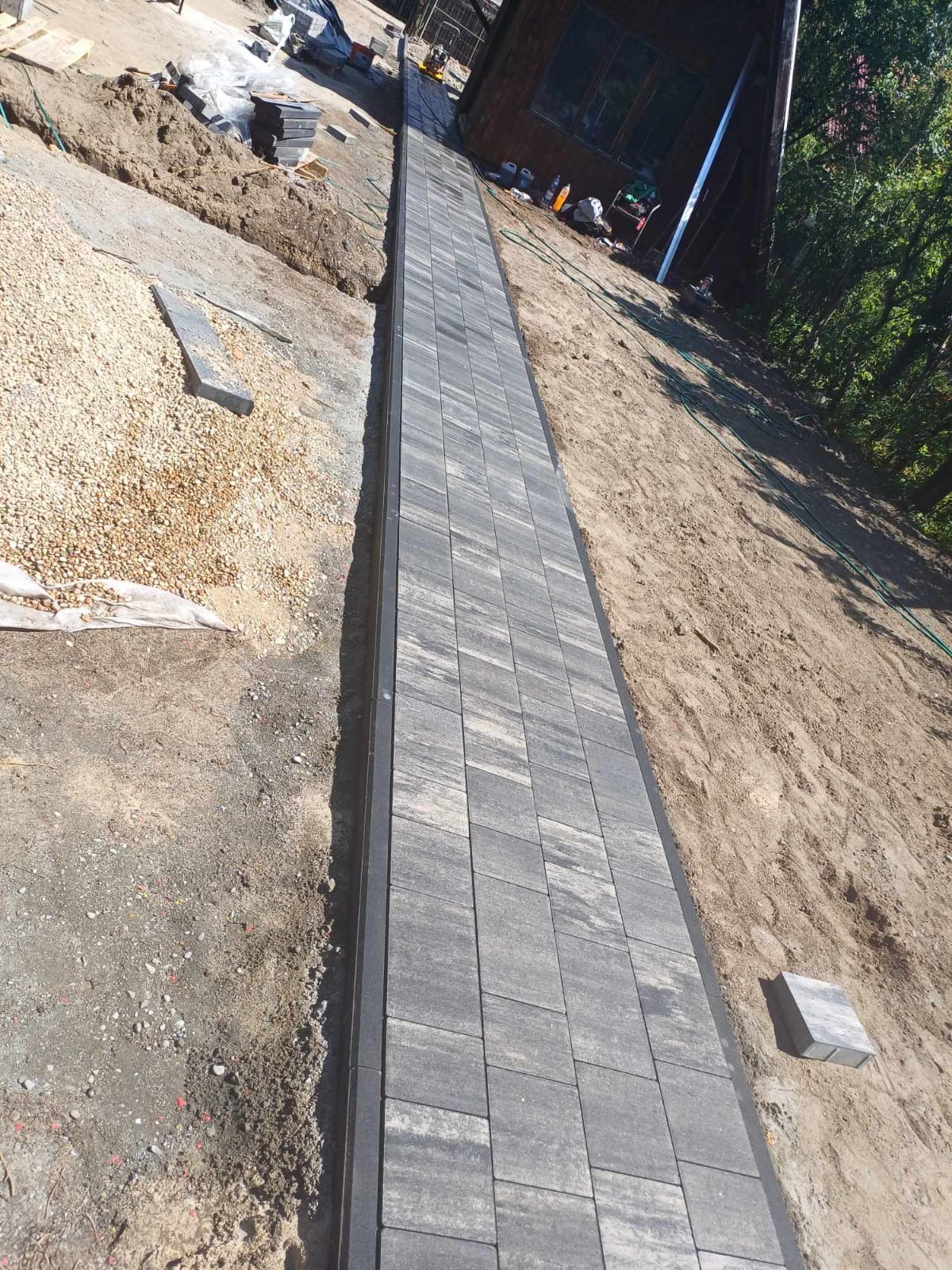 Układanie prostokątnej ścieżki z szarej kostki brukowej z czarnym obrzeżem w Tczewie, widoczny fragment przygotowanego podłoża i kostka do ułożenia.