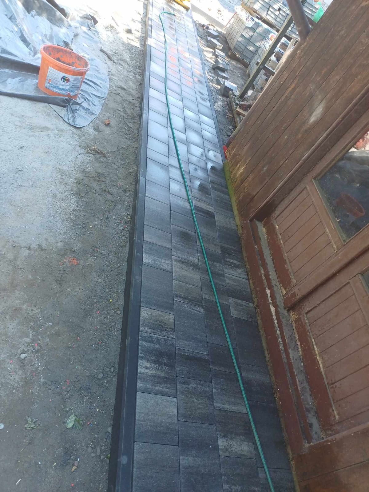 Ułożona ścieżka z szarej kostki brukowej prowadząca wzdłuż drewnianego budynku, widoczny pomarańczowy kubeł i zielony wąż ogrodowy.