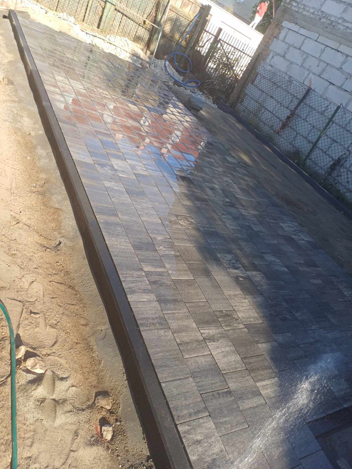 Świeżo ułożona, wilgotna kostka brukowa w odcieniach szarości, ułożona w karo, z widocznym obrzeżem i fragmentem węża ogrodowego.