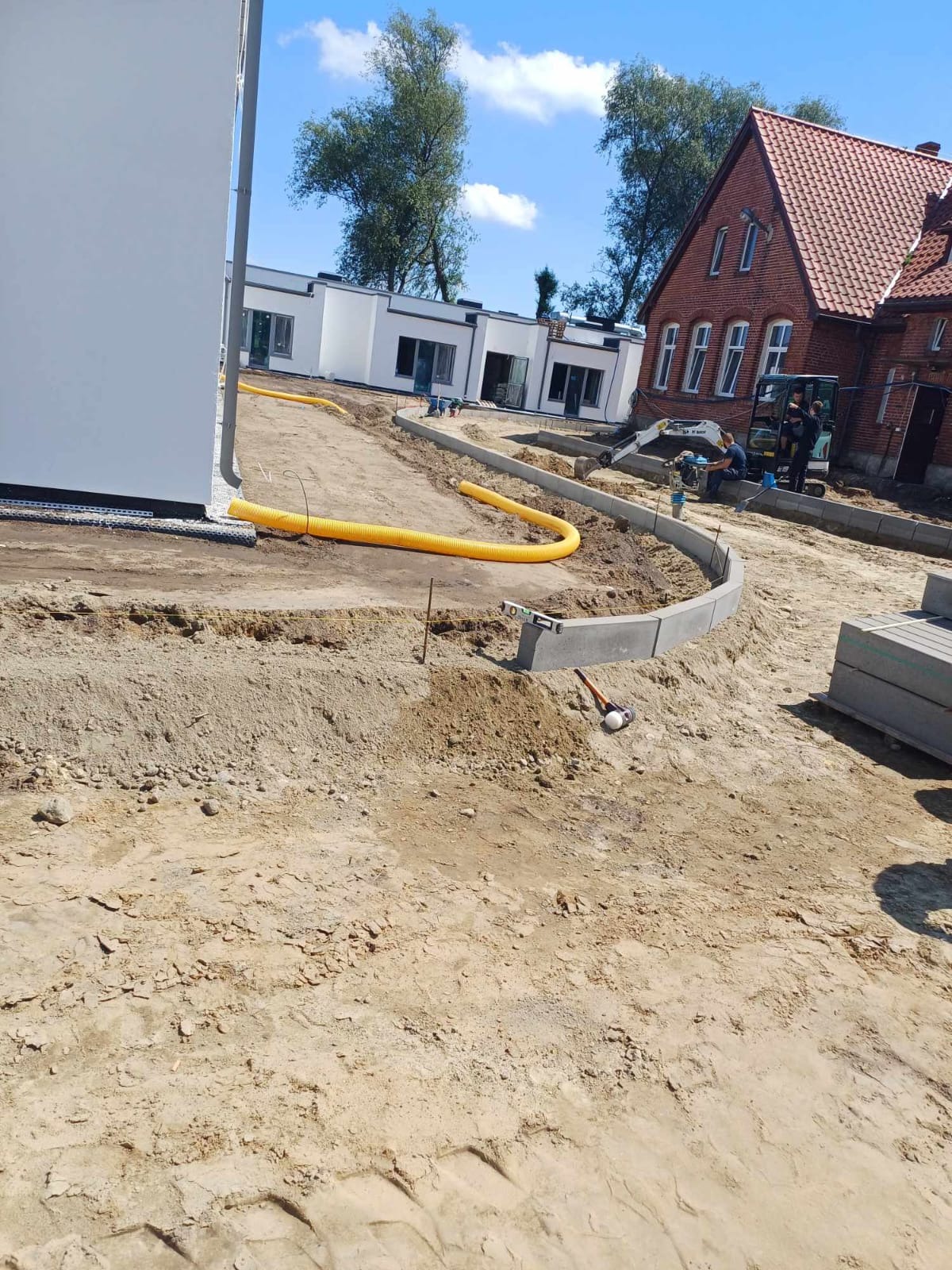 Przygotowanie terenu pod budowę chodnika z obrzeżami, widoczny minikoparka i żółty przewód, w tle budynki mieszkalne.