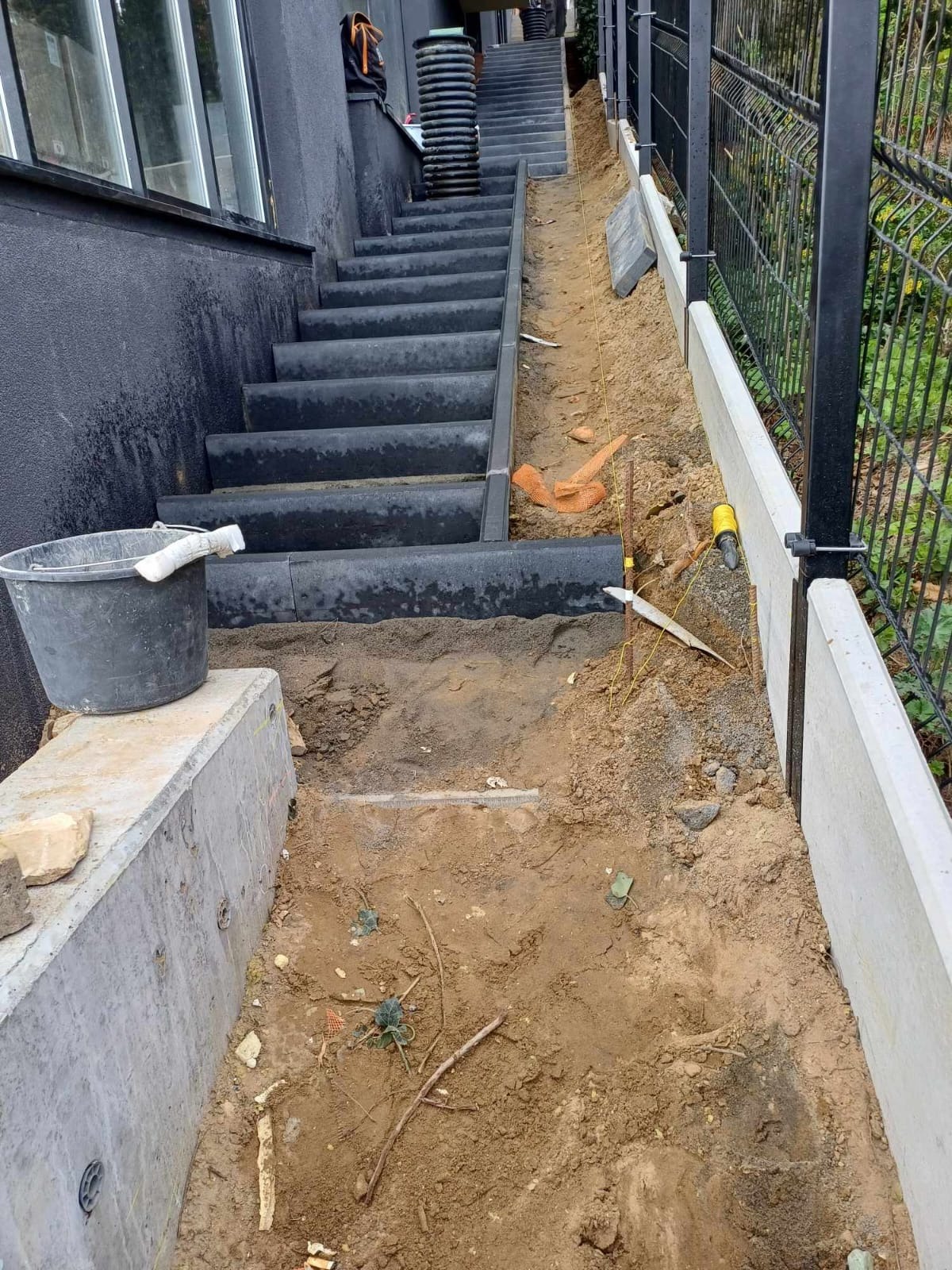 Przygotowanie terenu pod schody zewnętrzne z kostki, widoczne narzędzia, wykop i obrzeża betonowe wzdłuż ogrodzenia.
