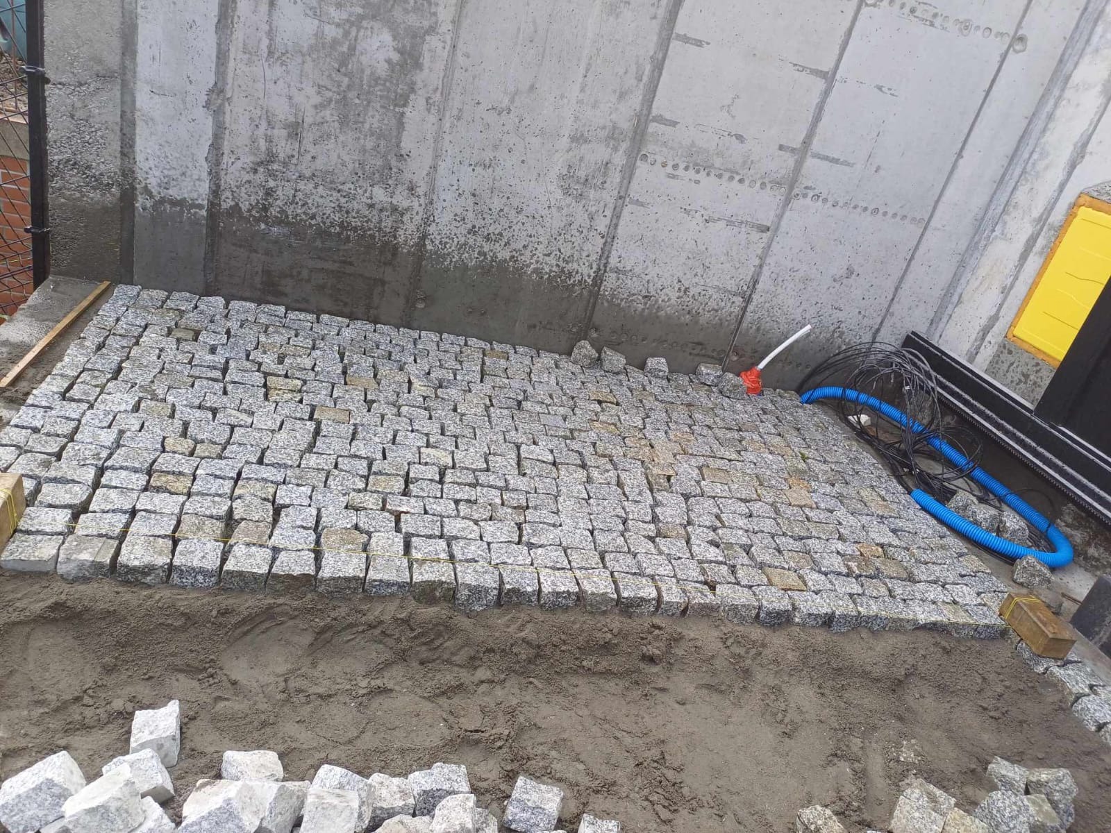 Fragment układanej nawierzchni z kostki granitowej, widoczne kable i rury instalacyjne, surowy betonowy mur w tle.