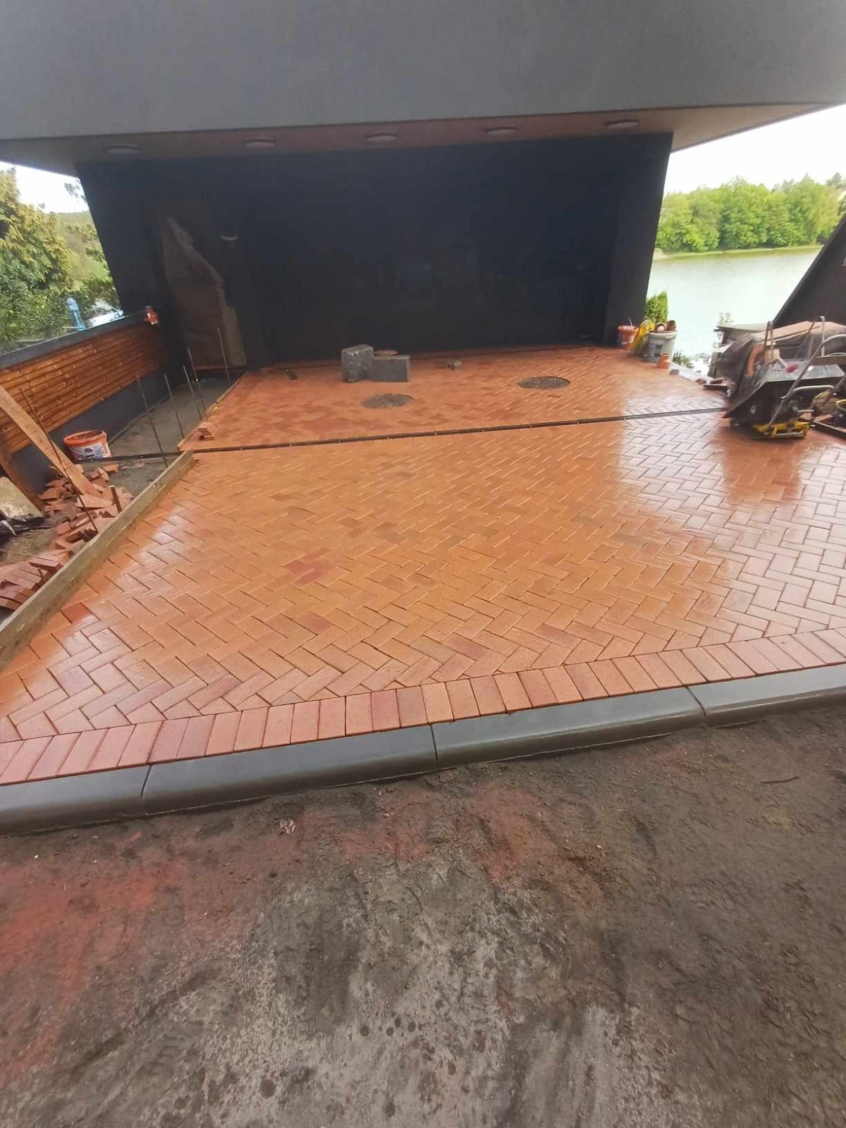 Czerwona kostka ułożona we wzór jodełki na wjeździe do garażu, z obrzeżem z szarej kostki, widoczny sprzęt budowlany i częściowo ułożona kostka po lewej stronie, w tle jezioro i zieleń.