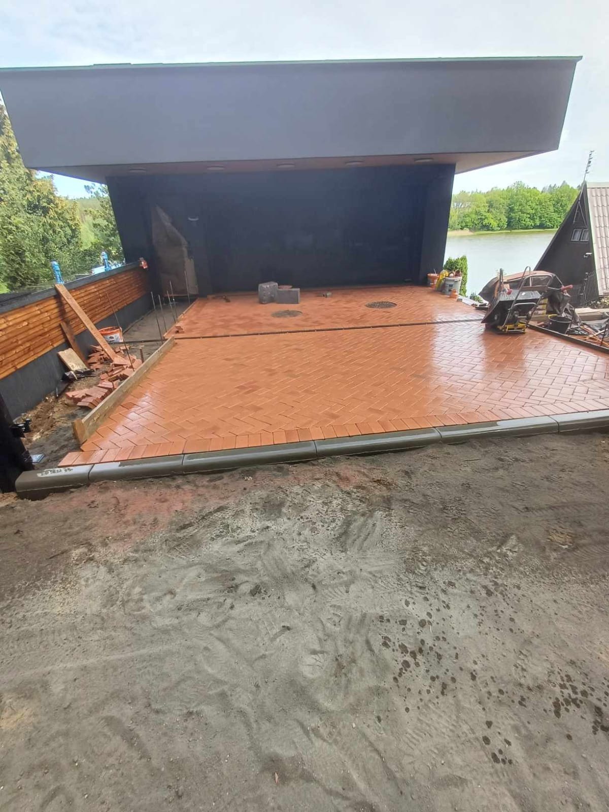 Układanie czerwonej kostki brukowej w jodełkę na posesji z widokiem na jezioro, widoczne narzędzia i niezakończone prace wykończeniowe.