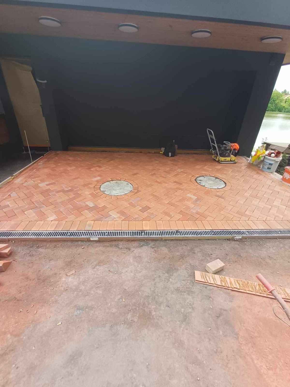 Nowa, czerwona kostka ułożona w jodełkę na tarasie z dwoma okrągłymi włazami i odpływem liniowym, widoczny zagęszczarka wibracyjna i jezioro w tle.