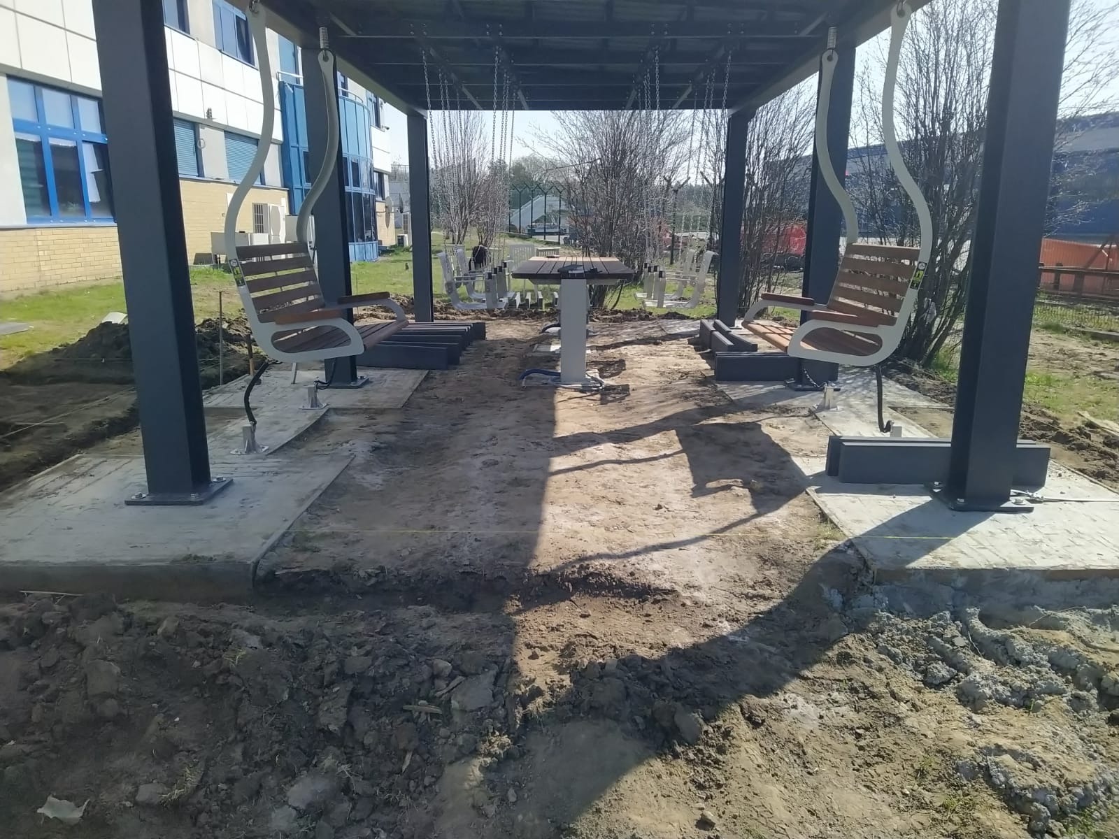 Montaż altany z huśtawkami: betonowe fundamenty, stalowe słupy, drewniane siedziska. Widoczne prace ziemne i przygotowanie terenu pod konstrukcję.