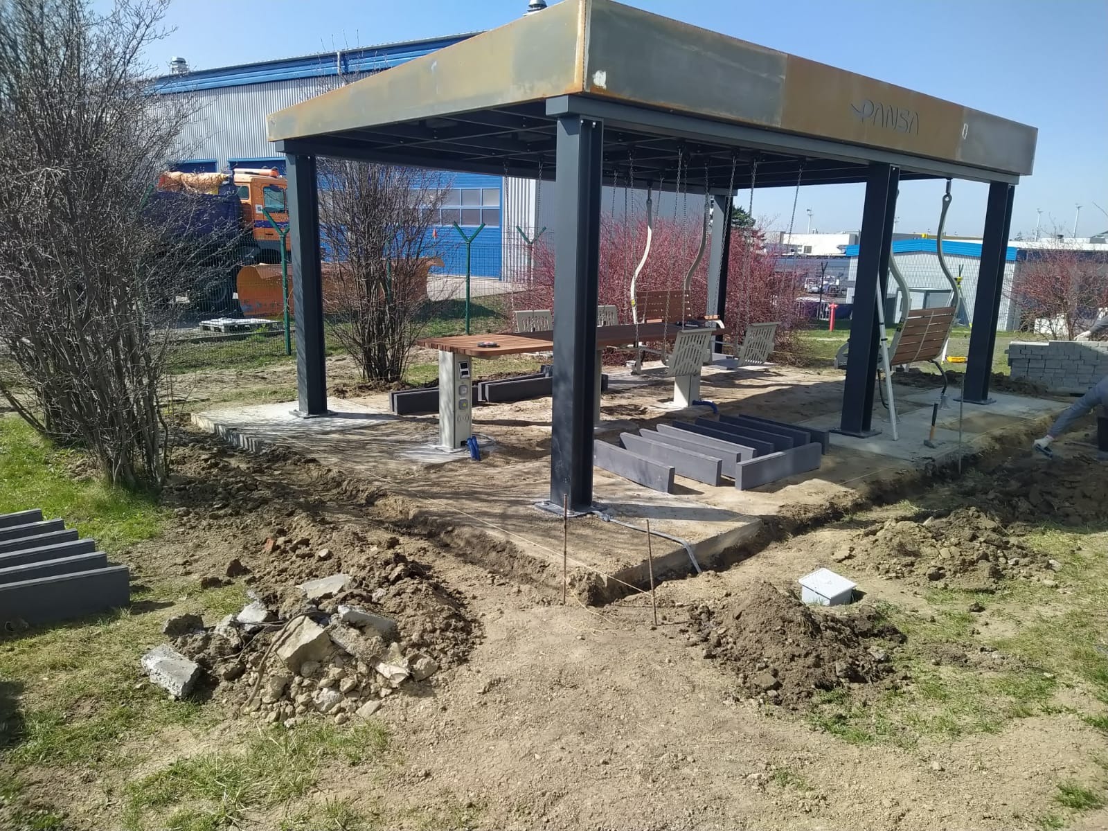 Budowa altany z huśtawkami i stołem na betonowej podstawie. Przygotowane obrzeża z kostki, teren częściowo rozkopany. W tle budynek przemysłowy i pojazd.