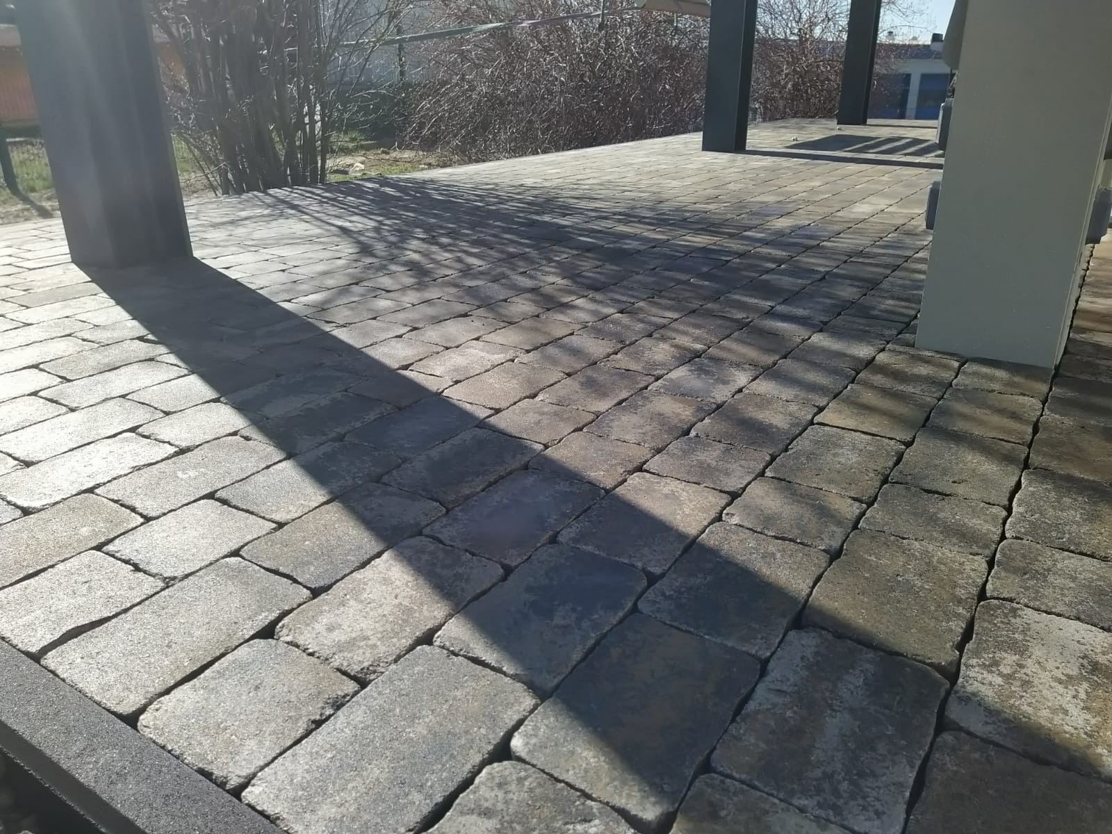 Ułożona kostka brukowa w odcieniach szarości tworzy wzór na zadaszonym tarasie. Cień słupa pada na kostkę, podkreślając jej fakturę i regularny układ.