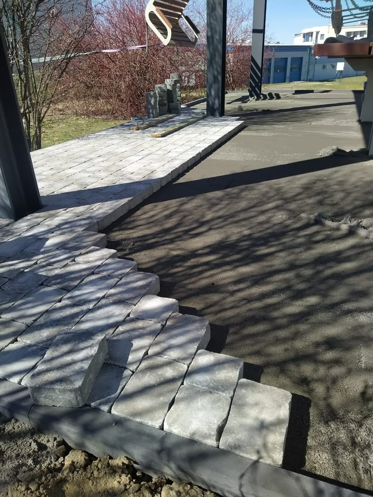 Fragment układania jasnoszarej kostki brukowej na podsypce. Częściowo ułożona nawierzchnia z kostki, widoczne zapasy kostki i przygotowane podłoże.