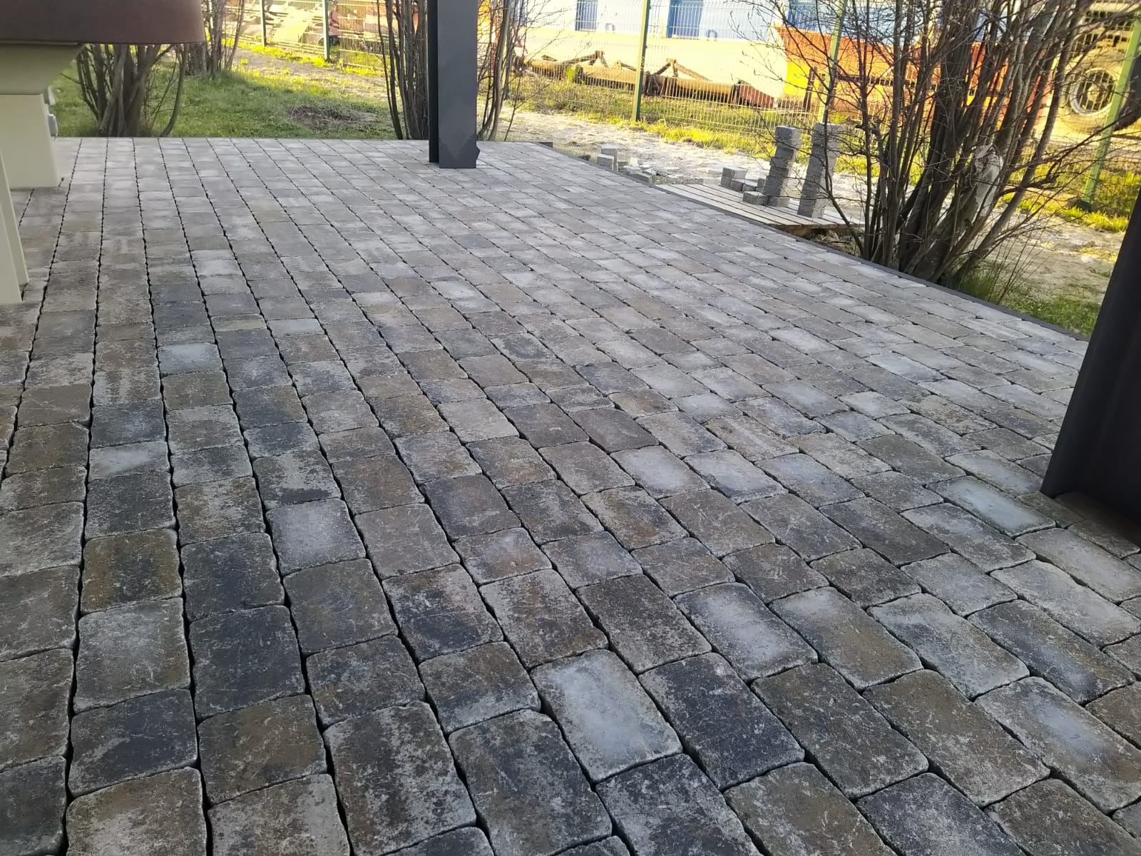 Ułożona, szara kostka brukowa na posesji, widok z perspektywy. W tle zieleń i elementy architektury. Kostka ułożona w regularny wzór, tworząc estetyczną powierzchnię.