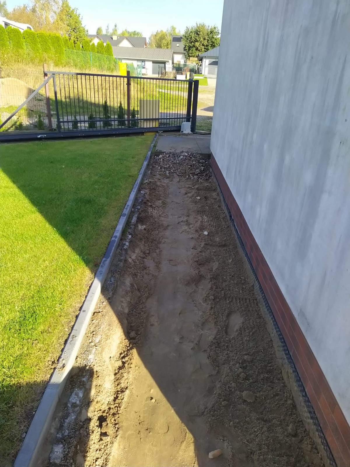 Przygotowany wykop pod ścieżkę obok domu z widocznymi obrzeżami i fragmentem trawnika. W tle brama wjazdowa i zieleń.