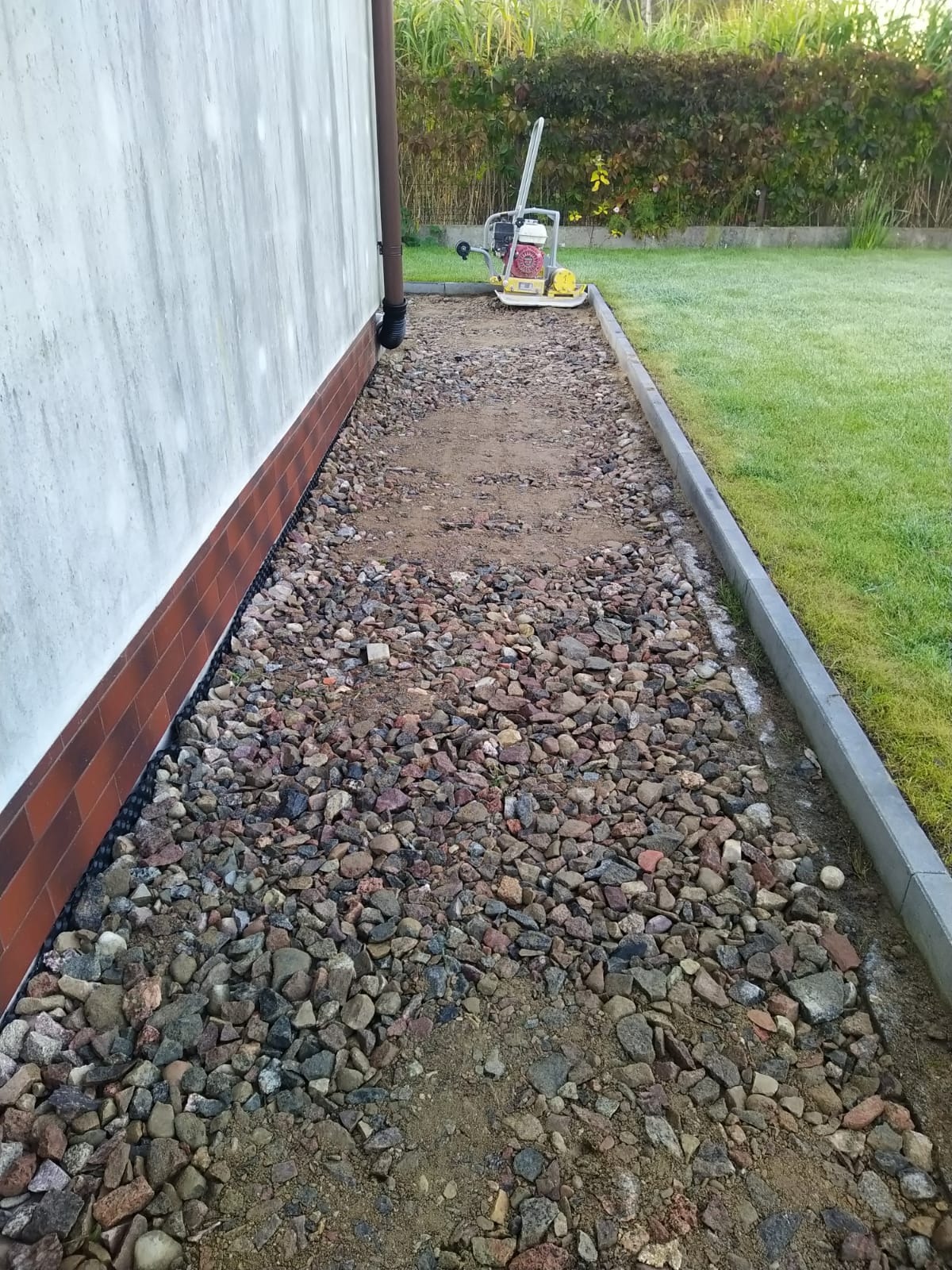 Przygotowanie podłoża pod ścieżkę z kamienia: podsypka z kamienia, obrzeża betonowe, ubijarka w tle, ściana budynku z rurą spustową. Widok z góry.