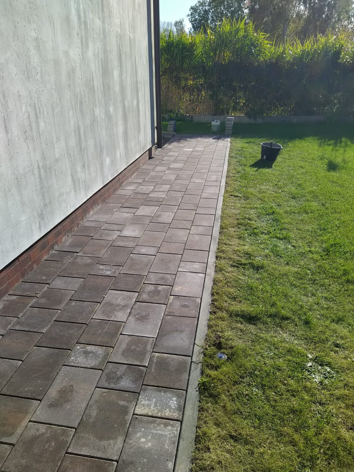 Ułożona ścieżka z kostki brukowej wzdłuż ściany budynku, z widocznym trawnikiem i zielenią w tle. Kostka w odcieniach szarości i brązu, regularny wzór.