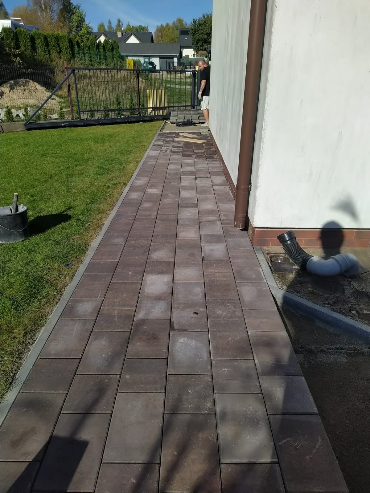 Ułożona ścieżka z kostki brukowej wzdłuż budynku, z widocznym systemem odprowadzania wody i fragmentem montowanego ogrodzenia w tle.