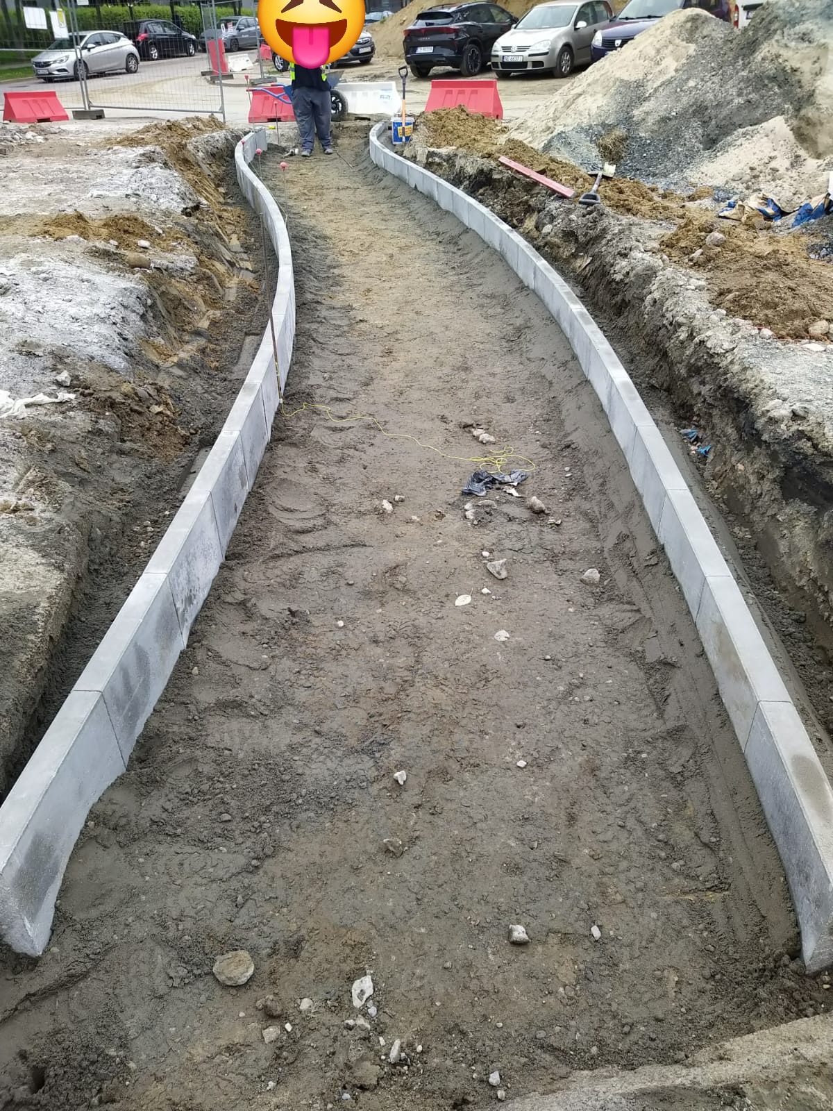 Zakrzywiony chodnik z szarej kostki obrzeżowej w trakcie budowy, widoczne ślady opon i ziemia. W tle zaparkowane samochody i osoba w odblaskowej kamizelce.