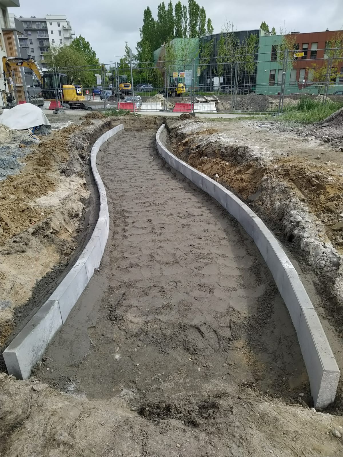 Wykopany, zakrzywiony chodnik z betonowymi obrzeżami w trakcie budowy. W tle budynki mieszkalne i maszyny budowlane. Widok z góry.