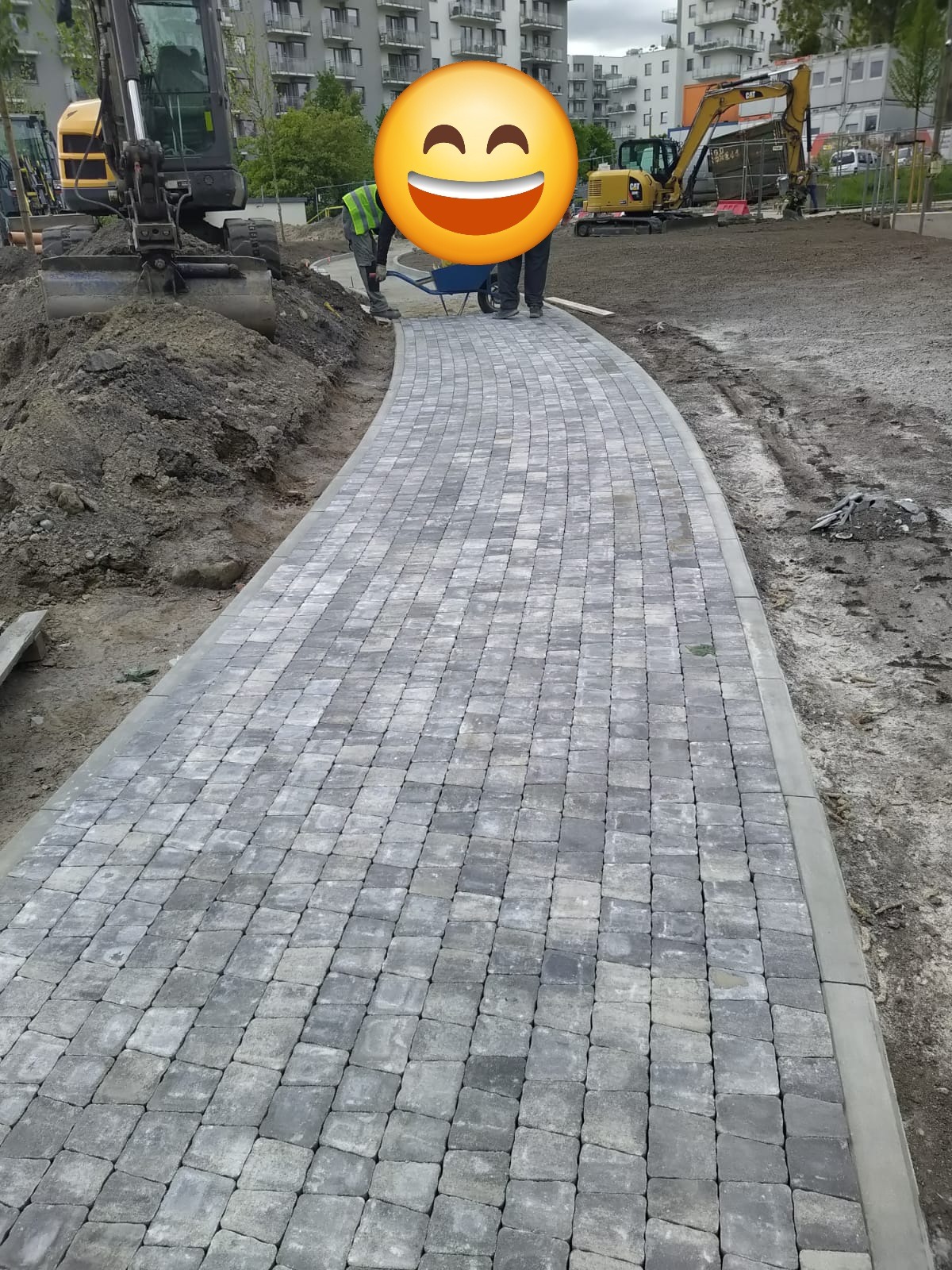 Ułożona, zakrzywiona ścieżka z szarej kostki brukowej, z widocznymi maszynami budowlanymi w tle. Etap prac w toku, teren przygotowany pod dalsze prace.