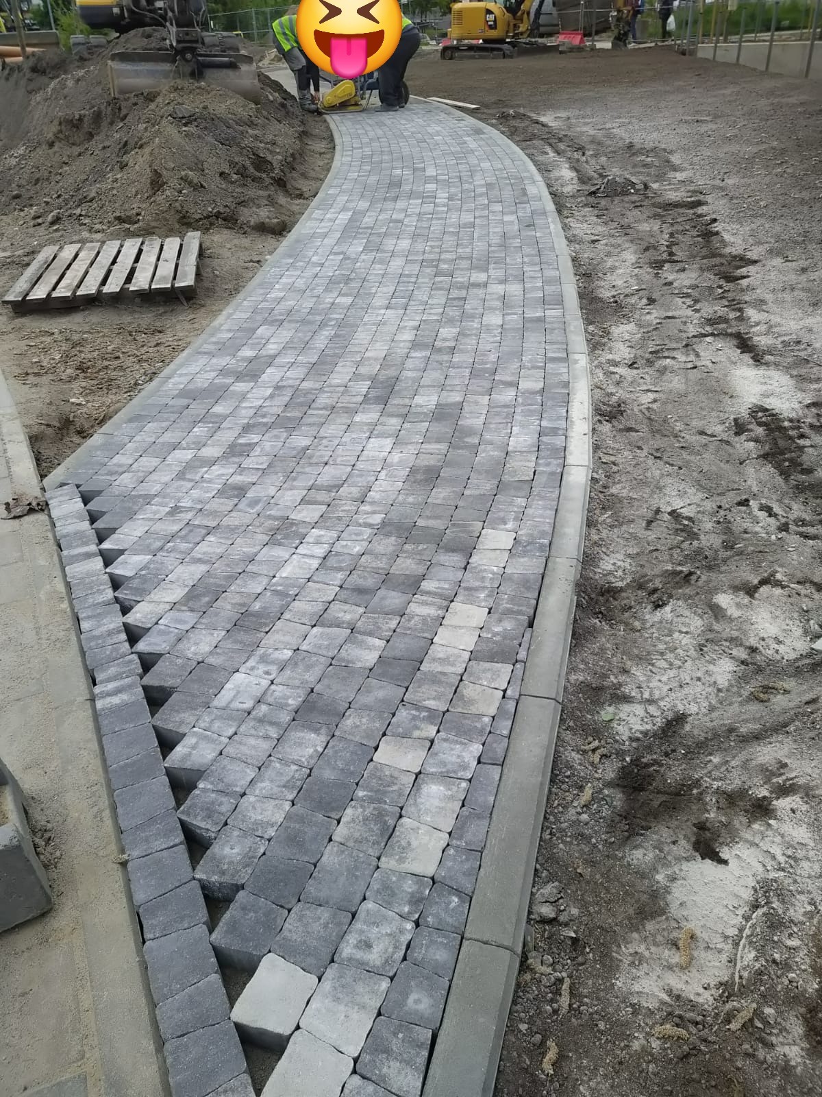 Zakrzywiony chodnik z szarej kostki brukowej ułożonej w romby, obrzeża betonowe. W tle maszyny budowlane i paleta. Częściowo ułożona nawierzchnia.