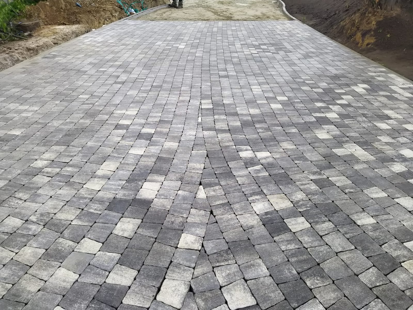 Ułożona kostka brukowa w odcieniach szarości, tworząca wzór zbiegający się w oddali. Widoczne krawężniki i fragmenty ziemi po bokach.