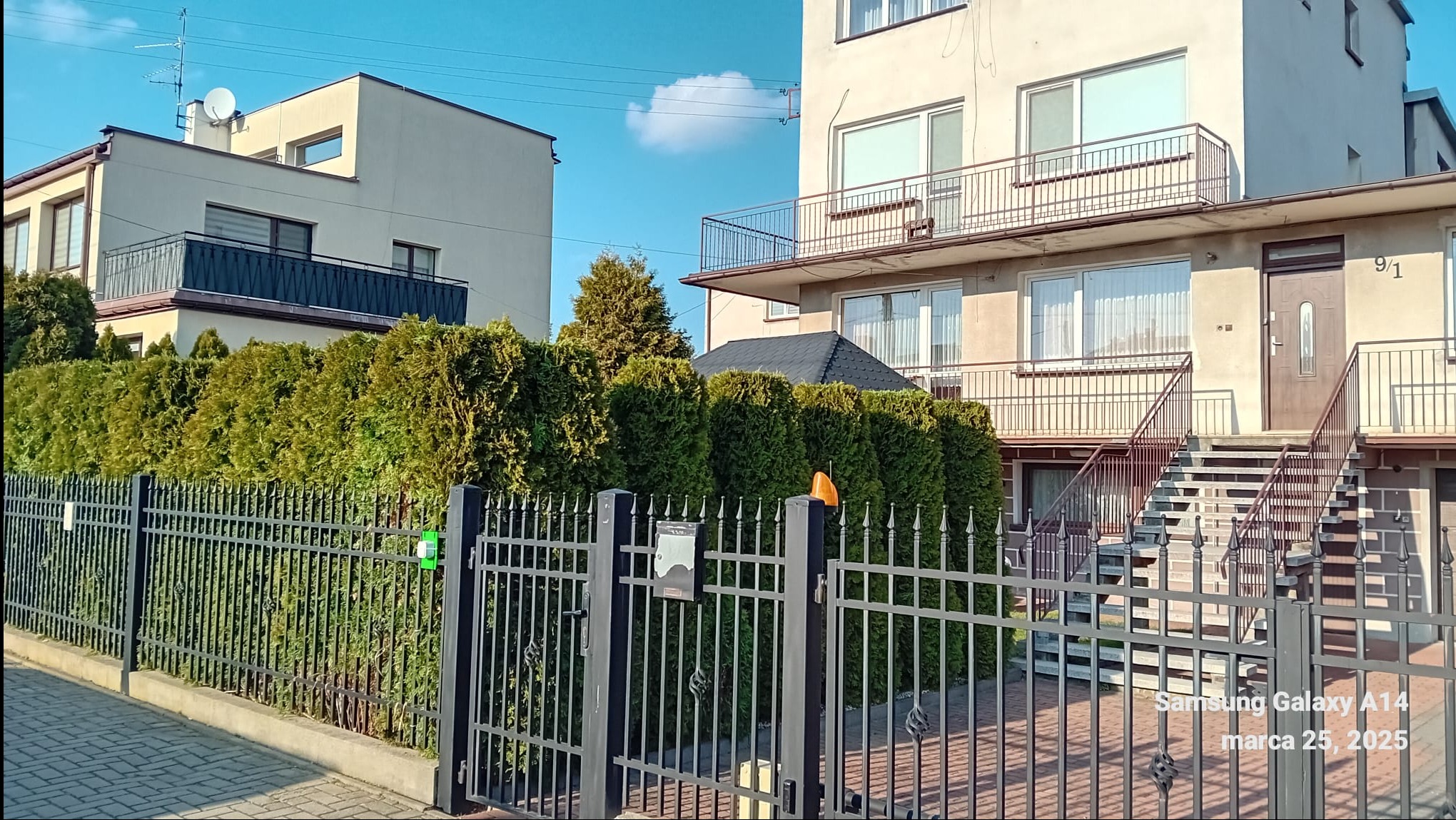 Dwupiętrowy dom z balkonami, ogrodzony metalowym płotem i żywopłotem z tuj, widok z ulicy w słoneczny dzień.