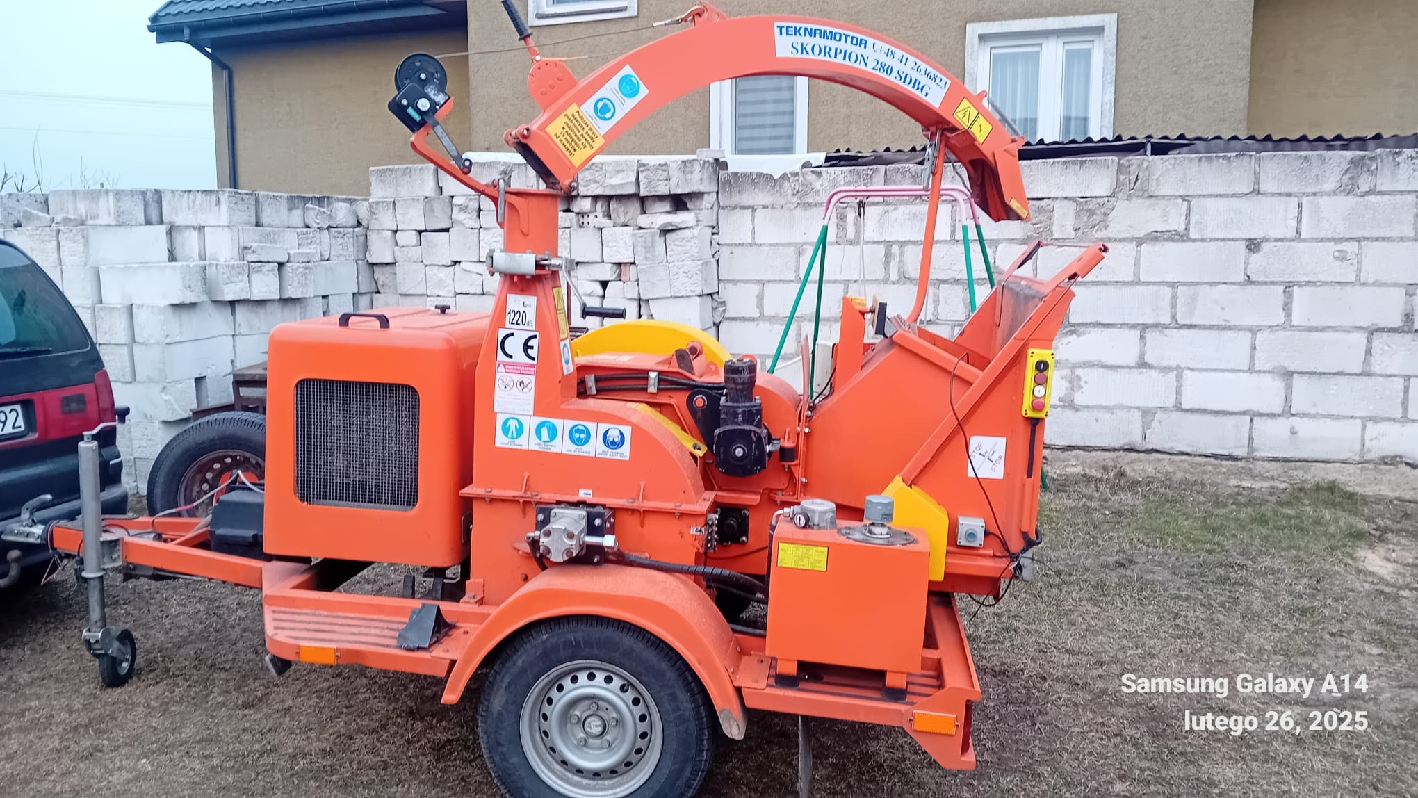 Pomarańczowa rębarka do gałęzi Teknamotor Skorpion 280 SDBG na przyczepie, zaparkowana na trawniku przed budynkiem z białego bloczka.