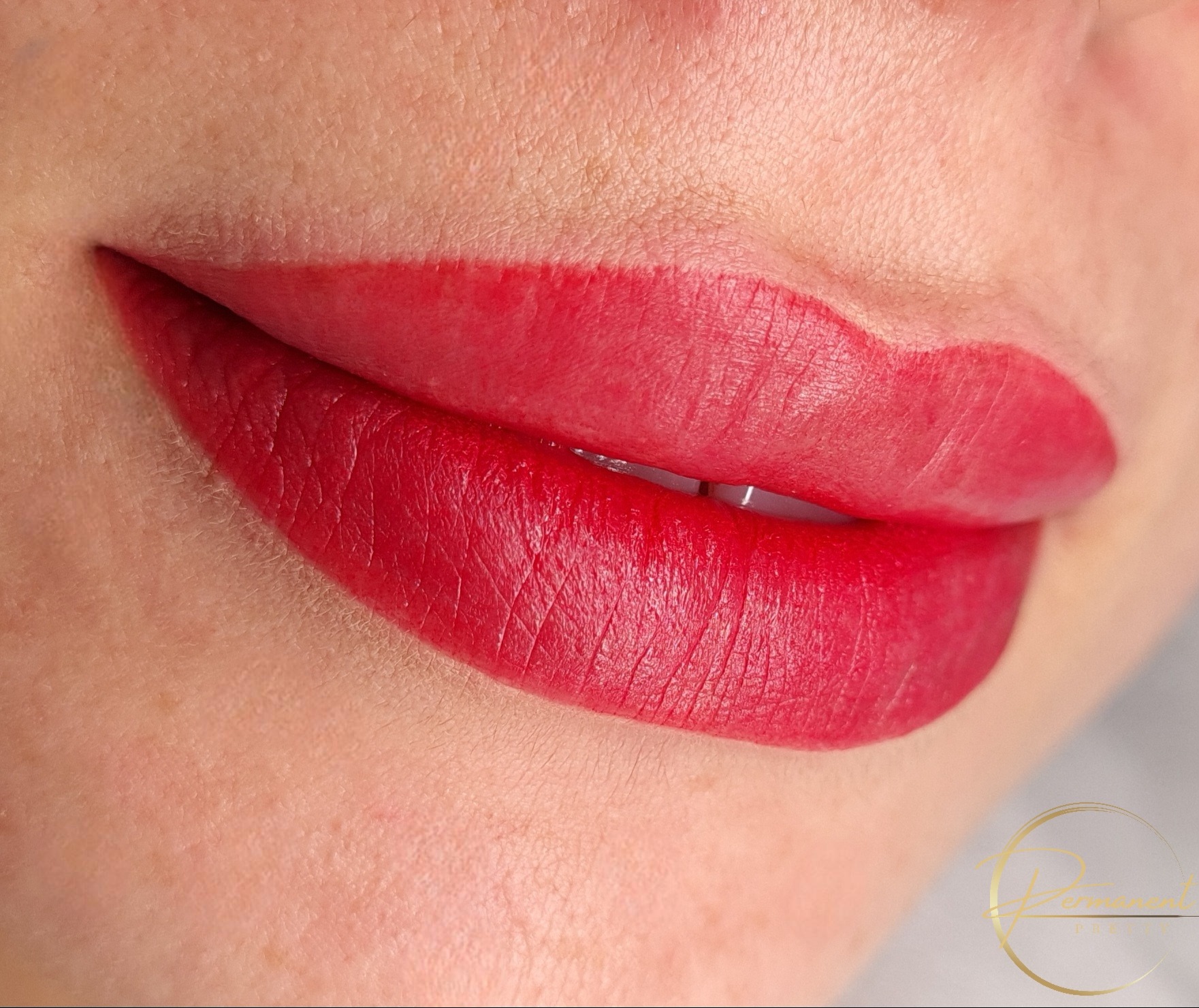 Zbliżenie na usta po zabiegu pigmentacji, intensywny czerwony kolor, widoczna struktura skóry i precyzyjny kontur, logo w prawym dolnym rogu.