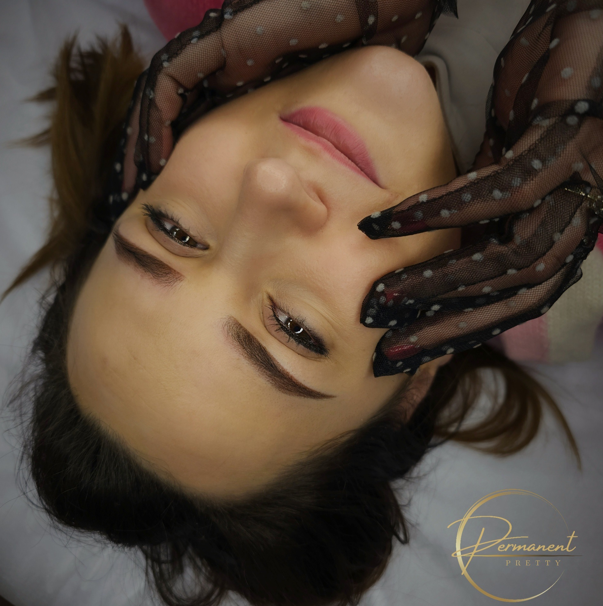 Kobieta leżąca na plecach z dłońmi w czarnych, koronkowych rękawiczkach z białymi kropkami, prezentująca efekt makijażu permanentnego brwi i delikatnego makijażu oczu.