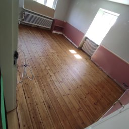 K.K.Cyklinowanie - Wnętrze pokoju z odświeżoną, drewnianą podłogą po cyklinowaniu, widoczne ślady po starej farbie na ścianach i grzejnikach, oraz kabel elektryczny zwisający z futryny.