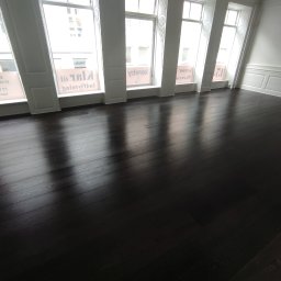 K.K.Cyklinowanie - Świeżo wycyklinowana, ciemna, drewniana podłoga w jasnym pokoju z wysokimi oknami i białymi ścianami z dekoracyjnymi panelami.