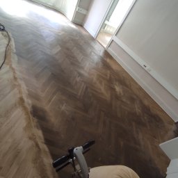 K.K.Cyklinowanie - Cyklinowanie parkietu ułożonego w jodełkę, widoczna maszyna do cyklinowania z workiem na pył, częściowo wycyklinowana podłoga w jasnym pomieszczeniu.