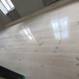 Wypolerowana, jasna podłoga drewniana z widocznymi słojami, odbijająca światło z okna, z listwami przypodłogowymi i grzejnikiem w tle.