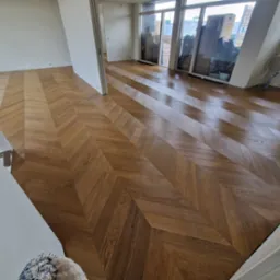 Nowoczesne wnętrze z parkietem ułożonym we wzór jodełki francuskiej, widok z perspektywy wejścia, duże okna z widokiem na miasto w tle.