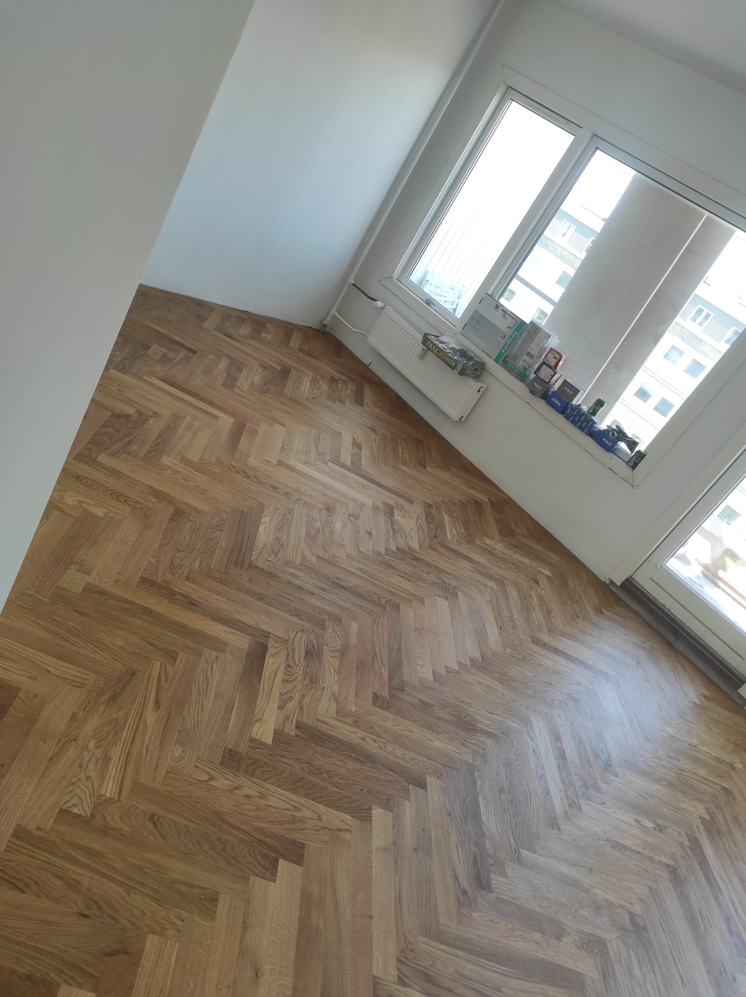 Ułożony parkiet w jodełkę w jasnym pokoju z białymi ścianami i oknem, na parapecie widoczne pudełka i opakowania różnych produktów.