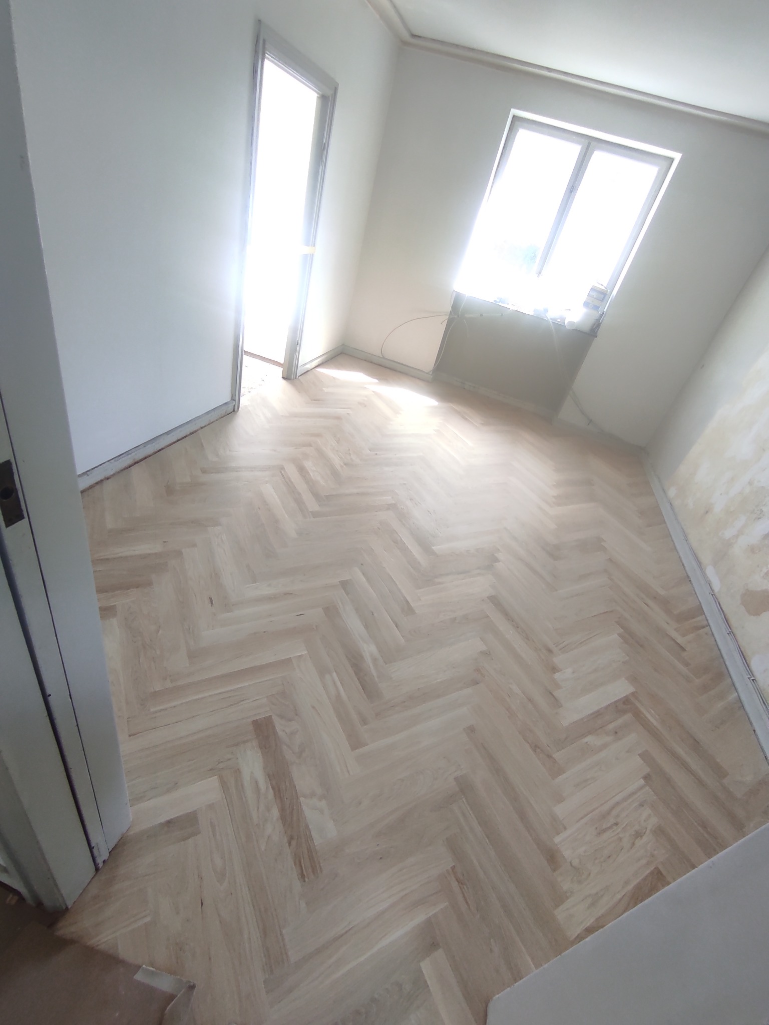 Jasny parkiet ułożony w jodełkę w pustym pokoju z oknem i otwartymi drzwiami, widoczne ślady po remoncie na ścianie.