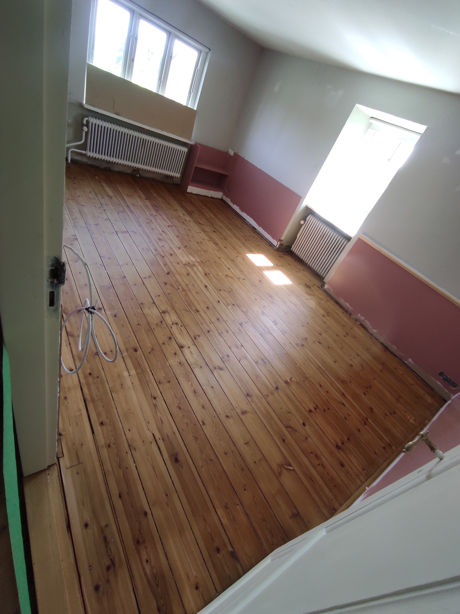 Wnętrze pokoju z odświeżoną, drewnianą podłogą po cyklinowaniu, widoczne ślady po starej farbie na ścianach i grzejnikach, oraz kabel elektryczny zwisający z futryny.