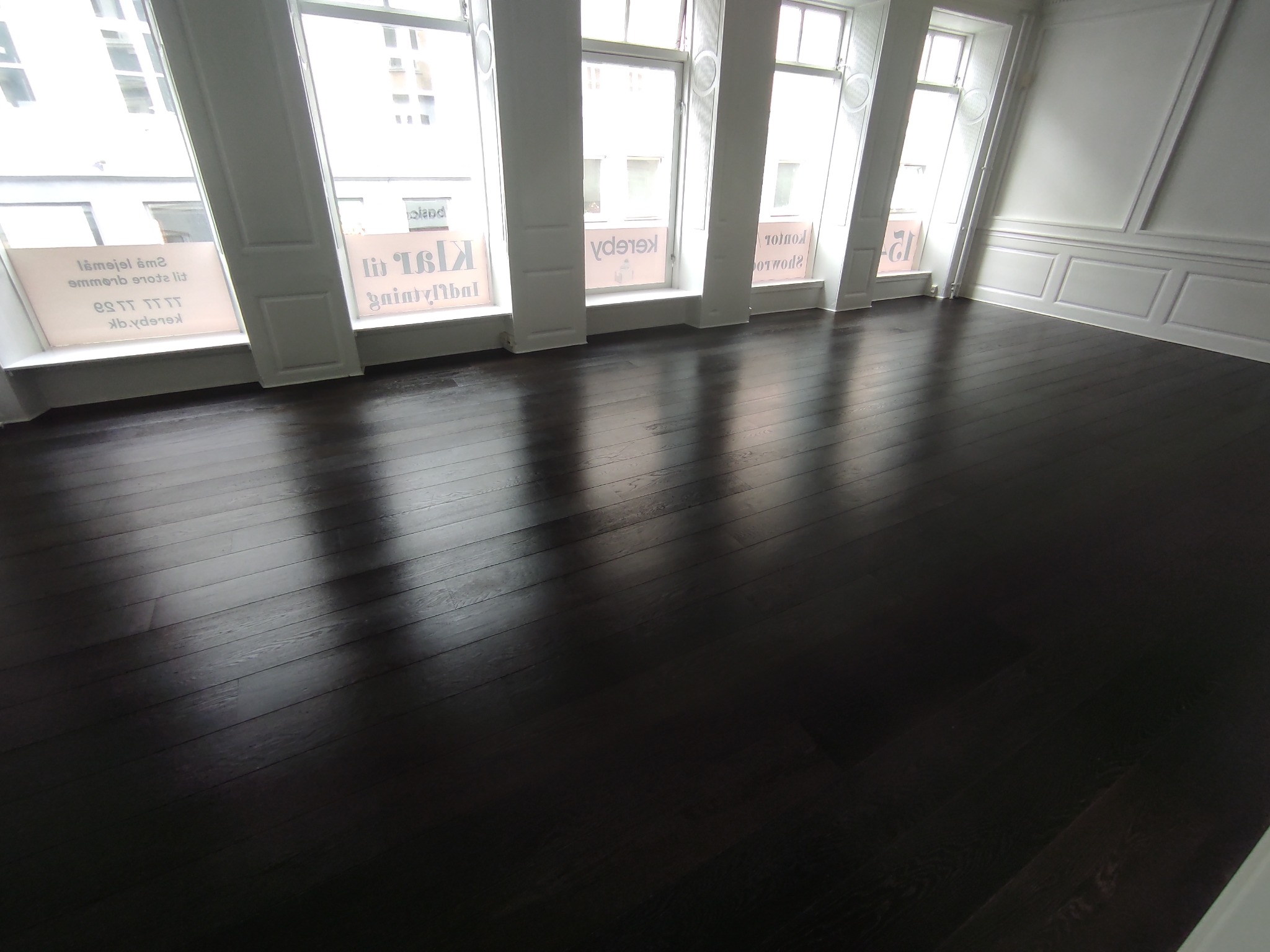 Świeżo wycyklinowana, ciemna, drewniana podłoga w jasnym pokoju z wysokimi oknami i białymi ścianami z dekoracyjnymi panelami.