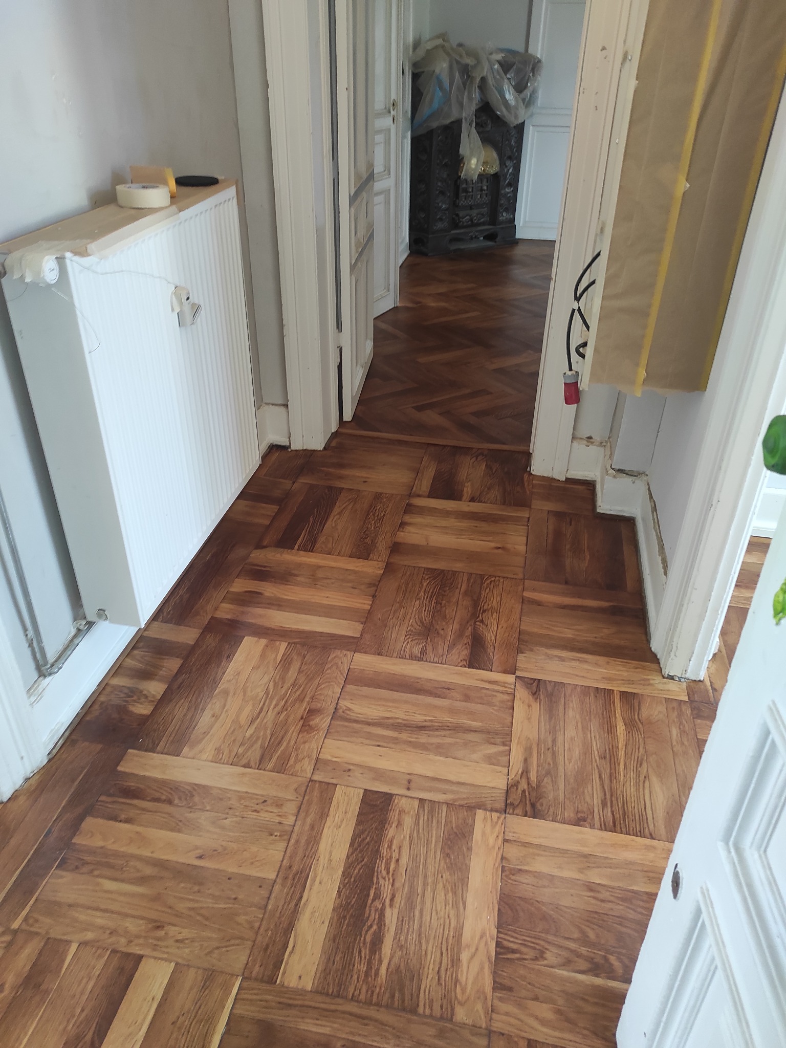 Ułożony parkiet w kwadraty w jasnym pomieszczeniu, widoczny fragment grzejnika z taśmą malarską i narzędziami, w tle parkiet ułożony w jodełkę w przejściu do kolejnego pomieszczenia.