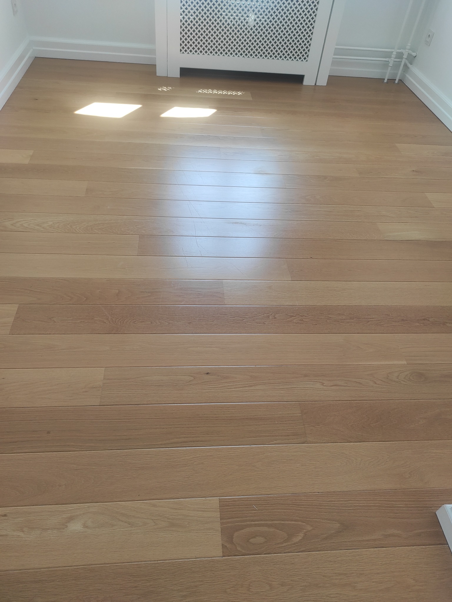 Jasna, drewniana podłoga z parkietu ułożona wzdłuż pokoju, widoczne listwy przypodłogowe i biały grzejnik z dekoracyjną osłoną, oświetlona przez promienie słoneczne wpadające przez okno.