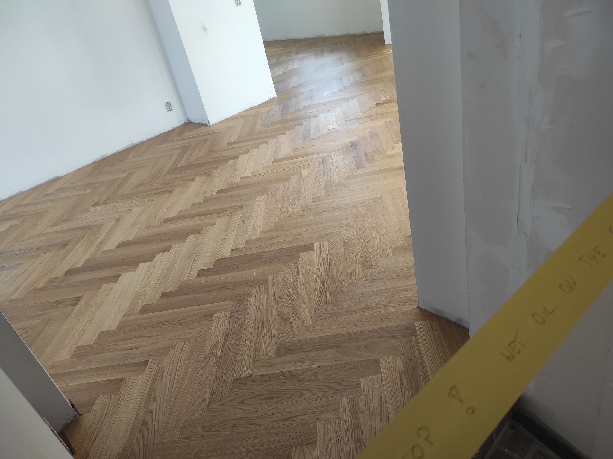 Ułożony parkiet w jodełkę, zabezpieczony żółtą taśmą z napisem 'Wet Oil on the ...', świeżo po olejowaniu, widok z góry.