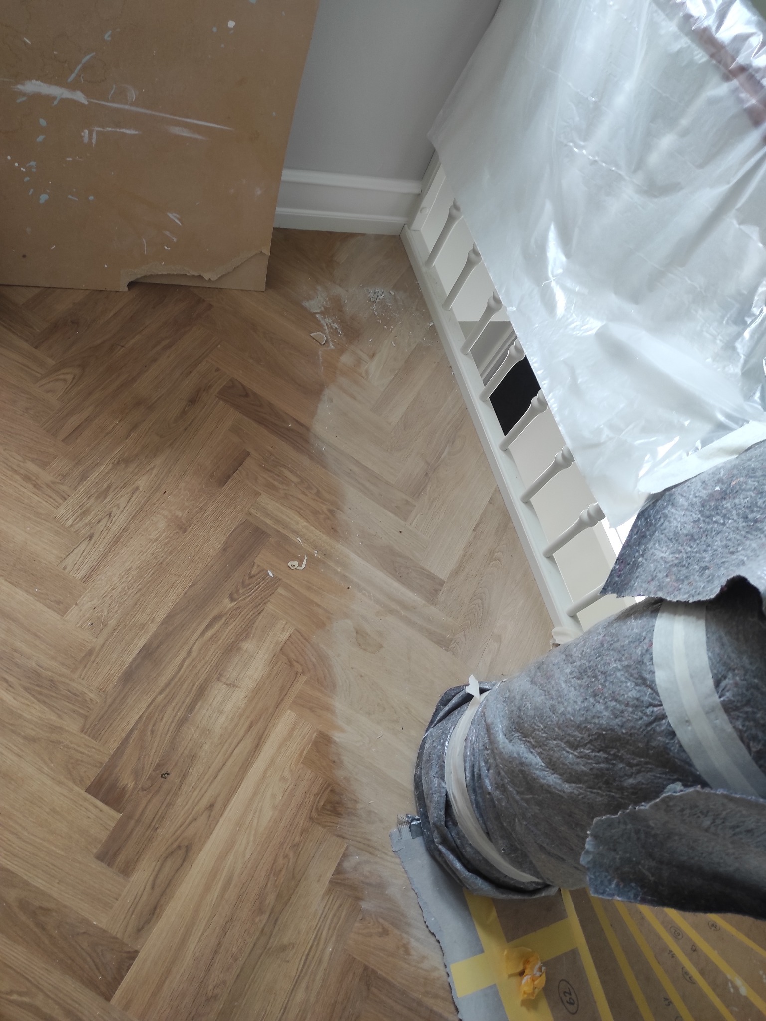 Podłoga ułożona w jodełkę, częściowo zakryta folią ochronną i rolką materiału zabezpieczającego, widoczne drobne zabrudzenia po remoncie.