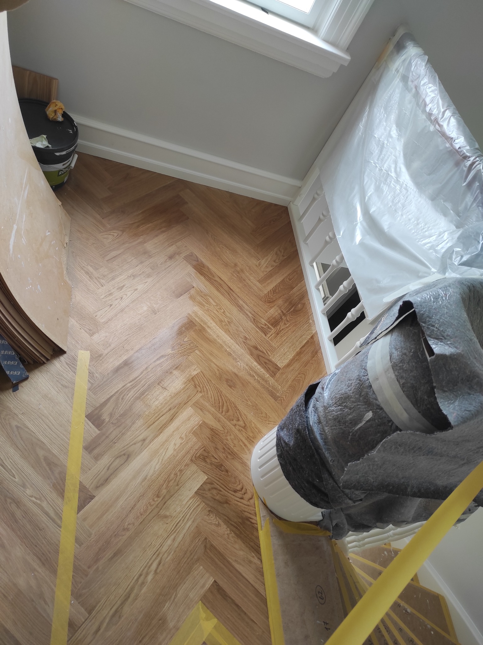 Ułożony parkiet w jodełkę, zabezpieczony taśmą malarską, z widocznym fragmentem schodów osłoniętych folią malarską i rolką materiału zabezpieczającego.