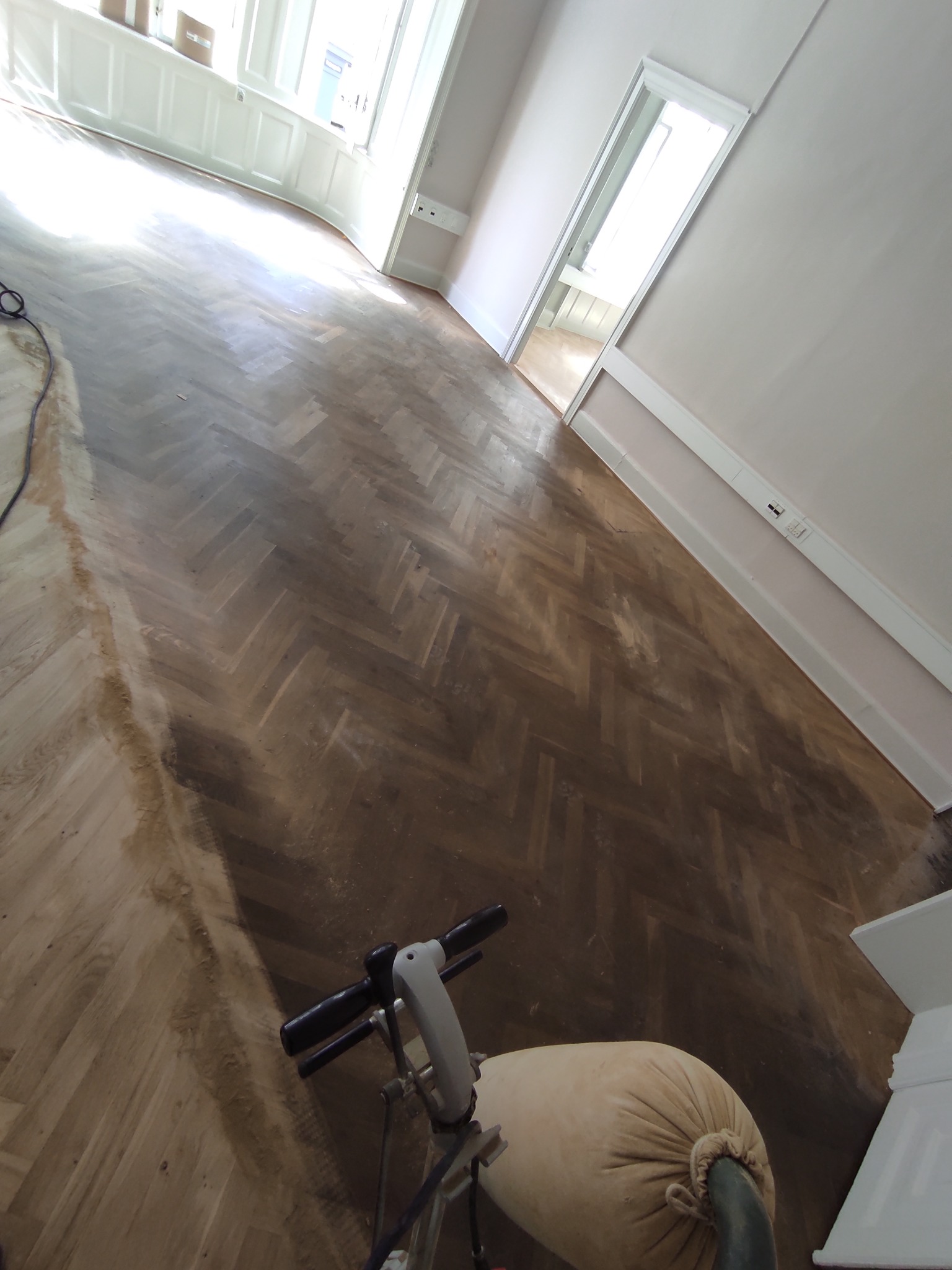 Cyklinowanie parkietu ułożonego w jodełkę, widoczna maszyna do cyklinowania z workiem na pył, częściowo wycyklinowana podłoga w jasnym pomieszczeniu.