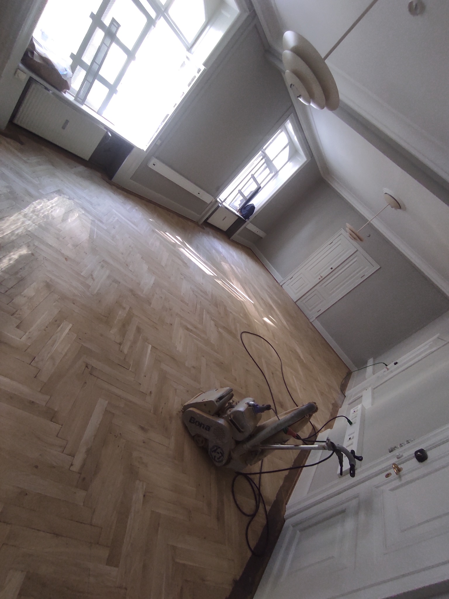 Maszyna Bona do obróbki parkietu w trakcie pracy, widoczne ślady po cyklinowaniu na parkiecie ułożonym w jodełkę, Kamionka Mała. W tle okno z jasnym światłem.