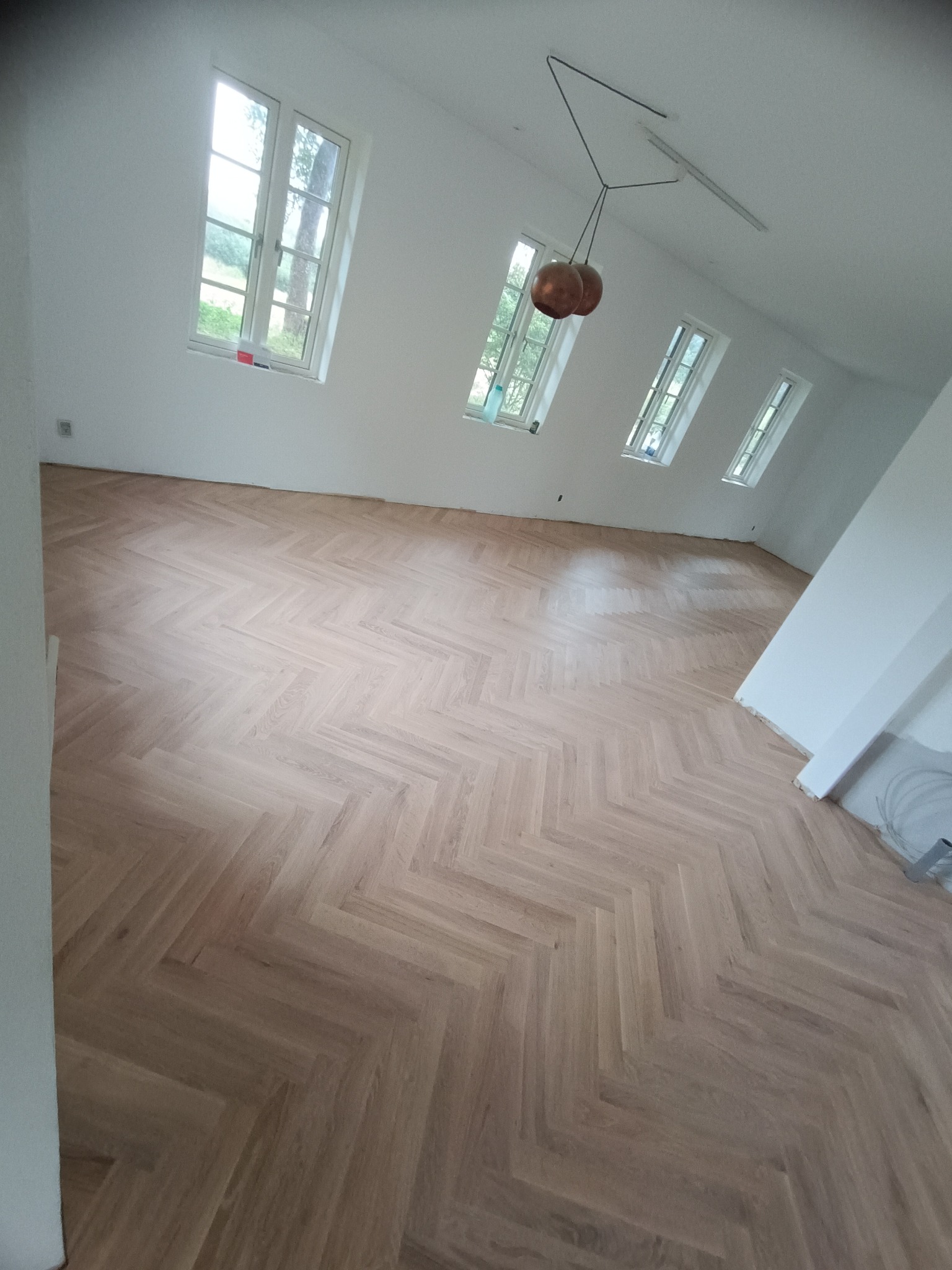 Jasny parkiet ułożony we wzór jodełki francuskiej w przestronnym, jasnym pokoju z kilkoma oknami i nowoczesną lampą wiszącą.