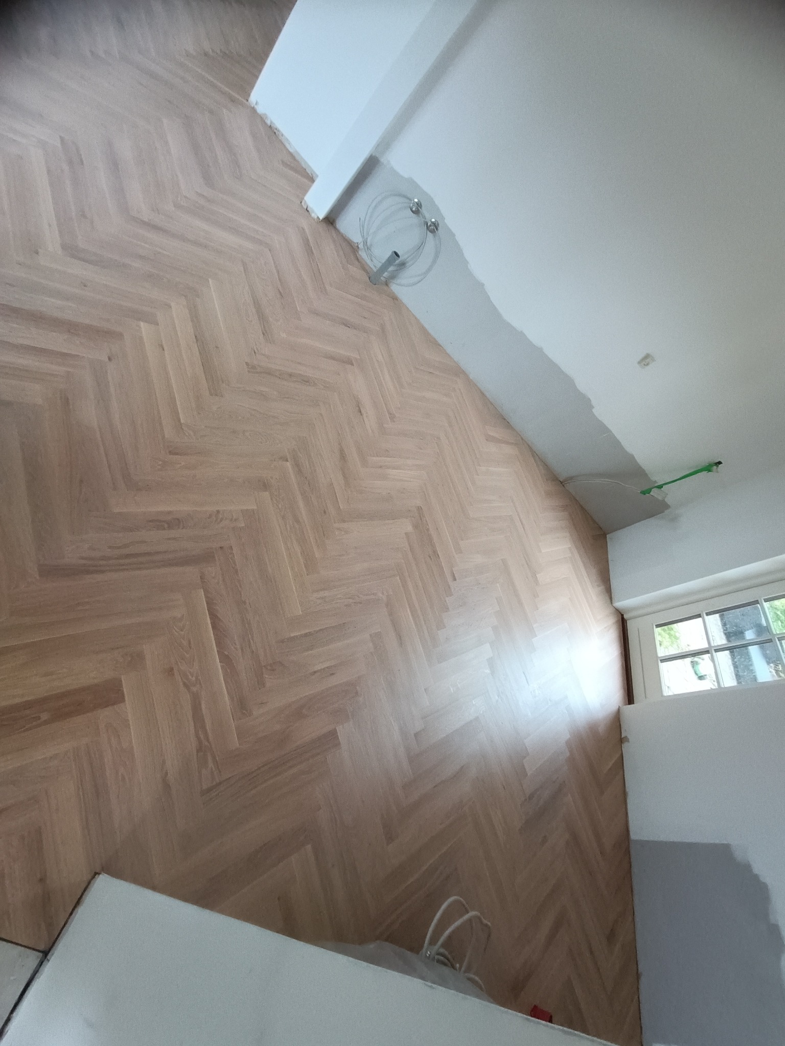 Ułożony parkiet w jodełkę, widoczne świeże łączenie ściany z sufitem, okno w tle.