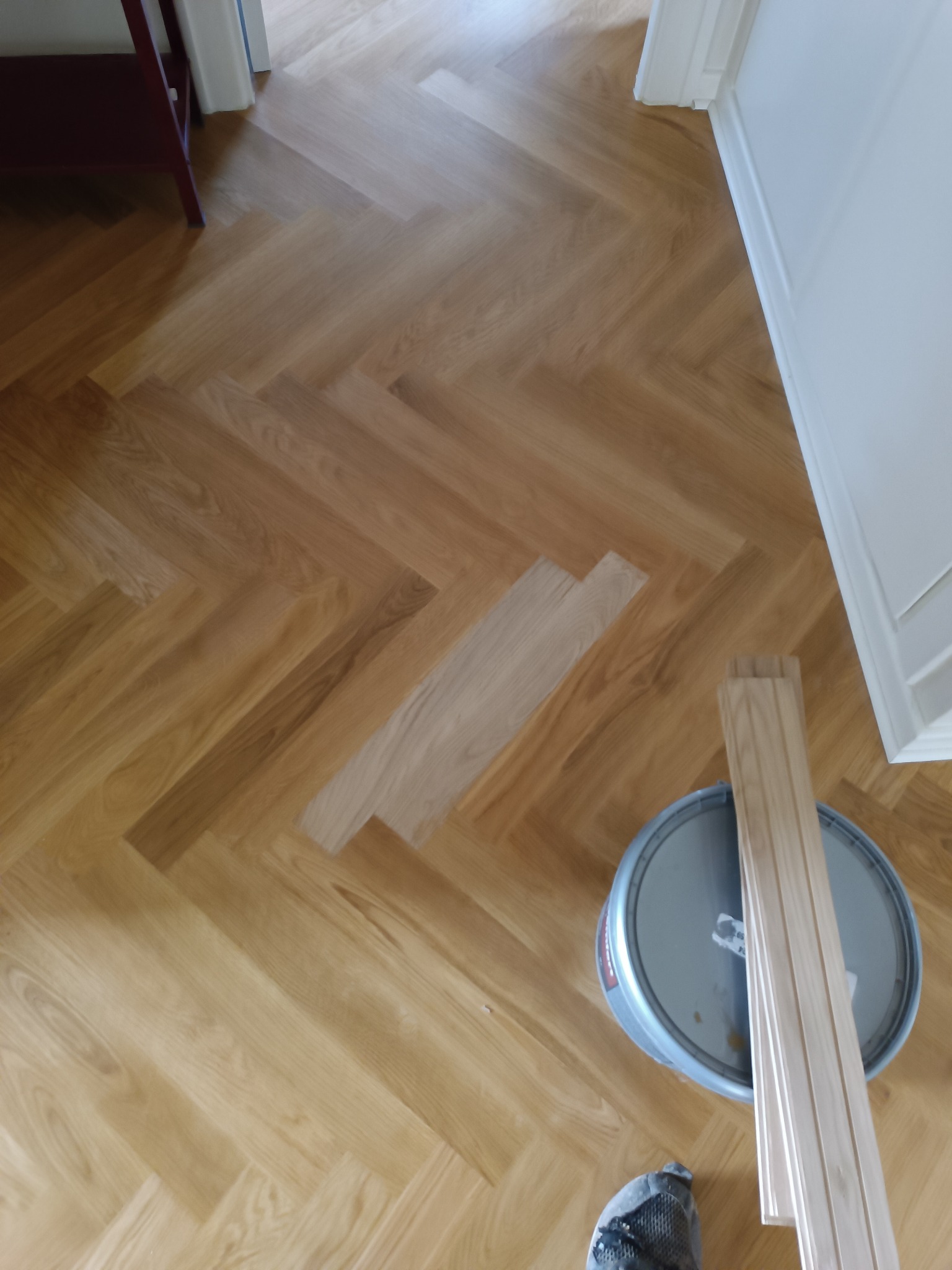 Ułożony parkiet w jodełkę z widocznym fragmentem po wymianie desek, obok puszka z klejem i listwy przypodłogowe.