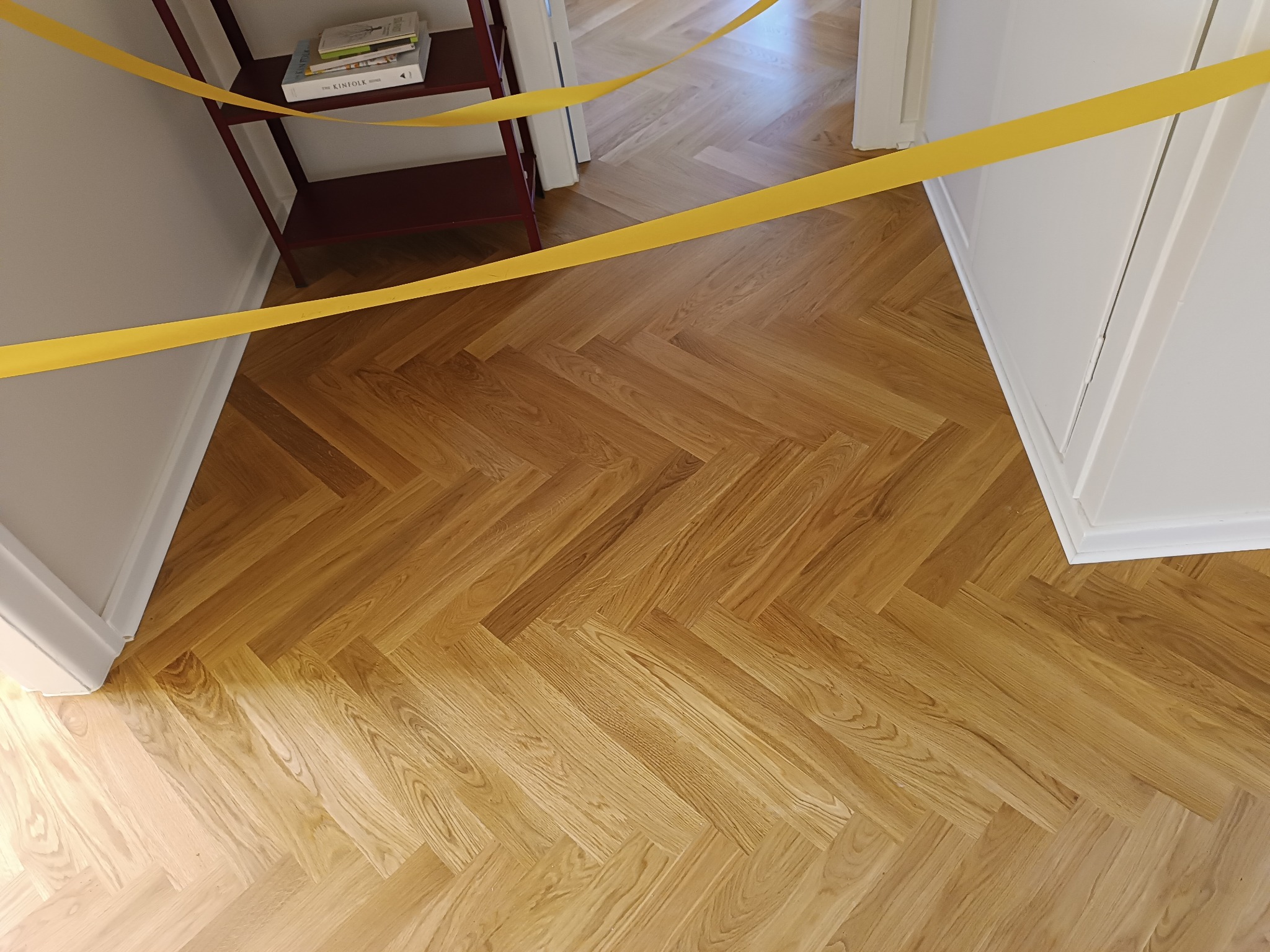 Nowo ułożony parkiet w jodełkę zabezpieczony żółtą taśmą ostrzegawczą, widoczny fragment regału z książkami.